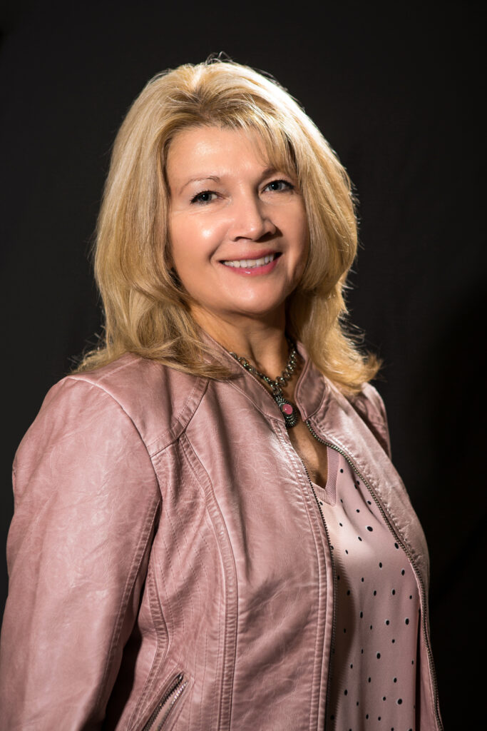 Portrait of a smiling woman in a pink jacket against a dark background.